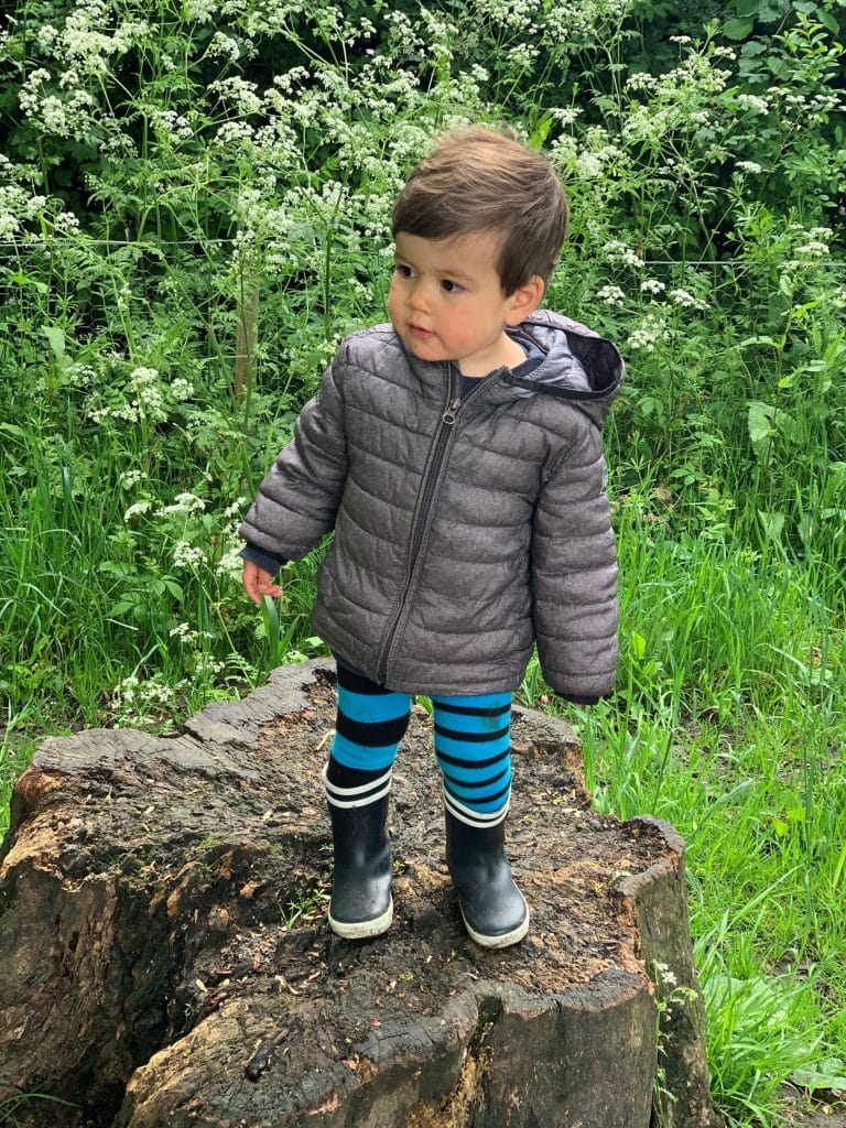 Patient after treatment in a gray jacket and striped leggings stands on a tree stump in a green, wooded area.