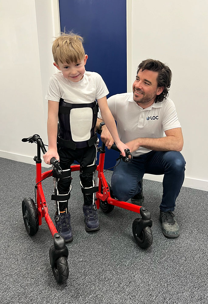 Ted stands in his custom Reciprocating Gait Orthosis with his walker, next to clinician Sam Walmsley, who made it for him